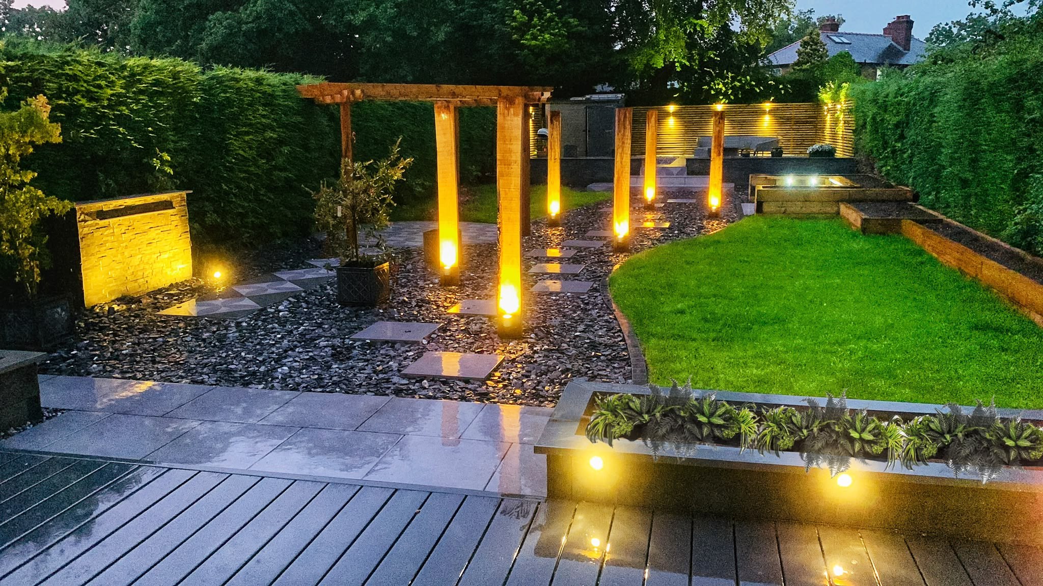 Dusk view of a garden with lit timber pergola posts lining a stone path, an illuminated water feature, grey composite decking and a striped lawn beyond a lit planter
