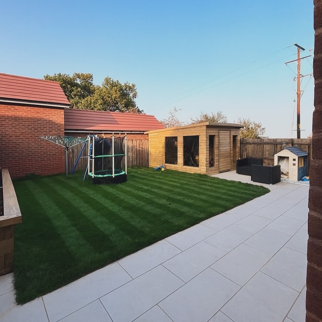 Striped lawn running beside a pale porcelain patio, with a timber garden room set against the boundary fence