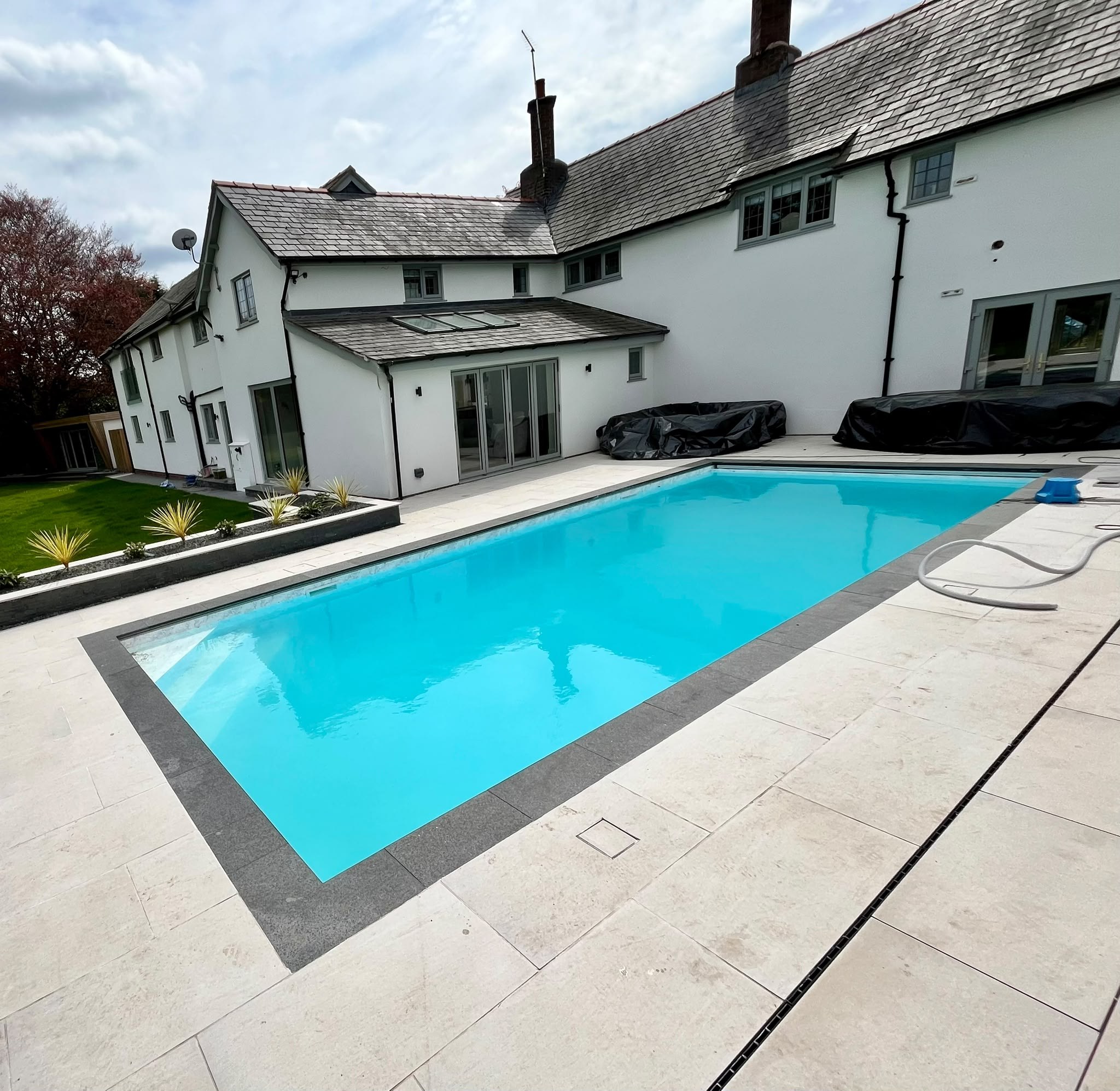 Rectangular swimming pool with dark stone coping and a pale stone surround beside a white rendered house
